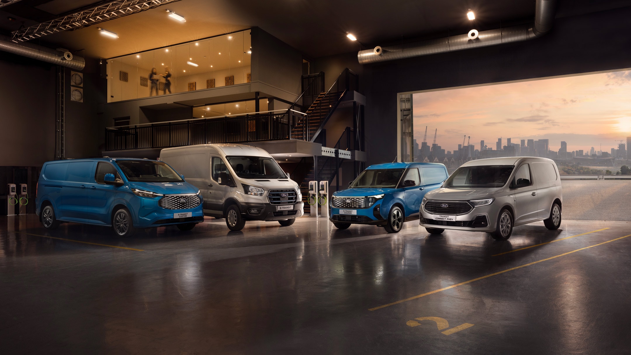 Sininen Ford E-Transit Custom, valkoinen Ford E-Transit, sininen Ford E-Transit Courier ja hopeinen Ford Transit Connect pysäköitynä moderniin autotalliin, josta näkyy kaupungin siluetti.