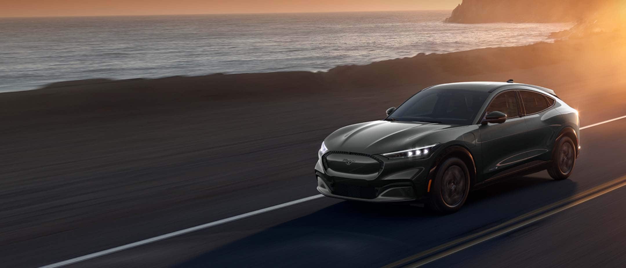 Tummanharmaa Ford Mustang Mach-E ajaa rannikkotiellä auringonlaskun aikaan.