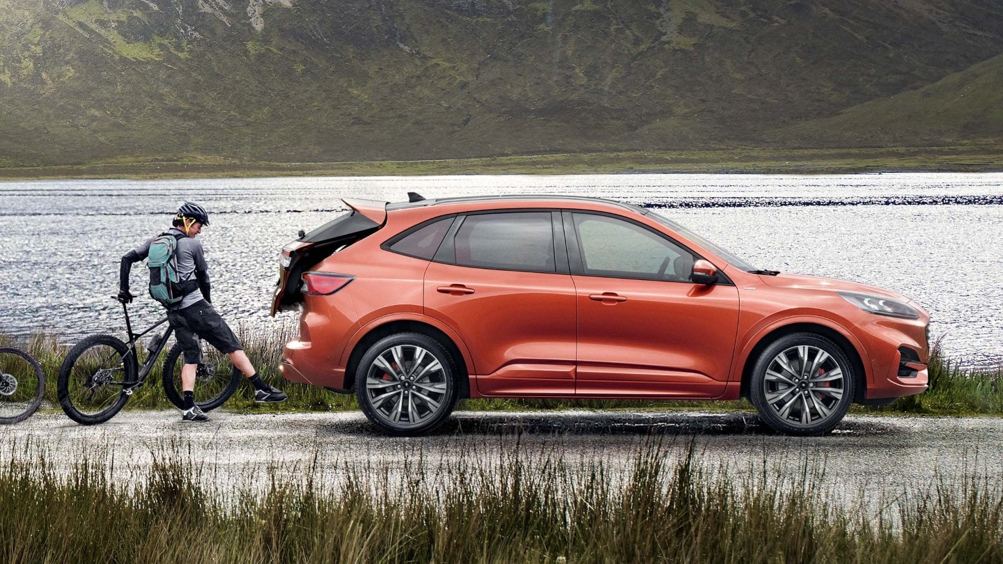 Oranssi Ford Kuga, jonka avoimeen tavaratilaan lastataan polkupyörää. Henkilö seisoo pyörän vieressä.