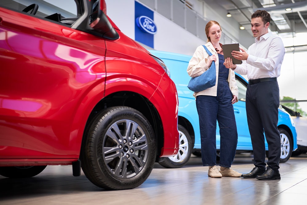 Kaksi ihmistä Ford-näyttelytilassa katsoo tablettia, etualalla punainen Ford-pakettiauto ja takana sininen Ford-pakettiauto. 