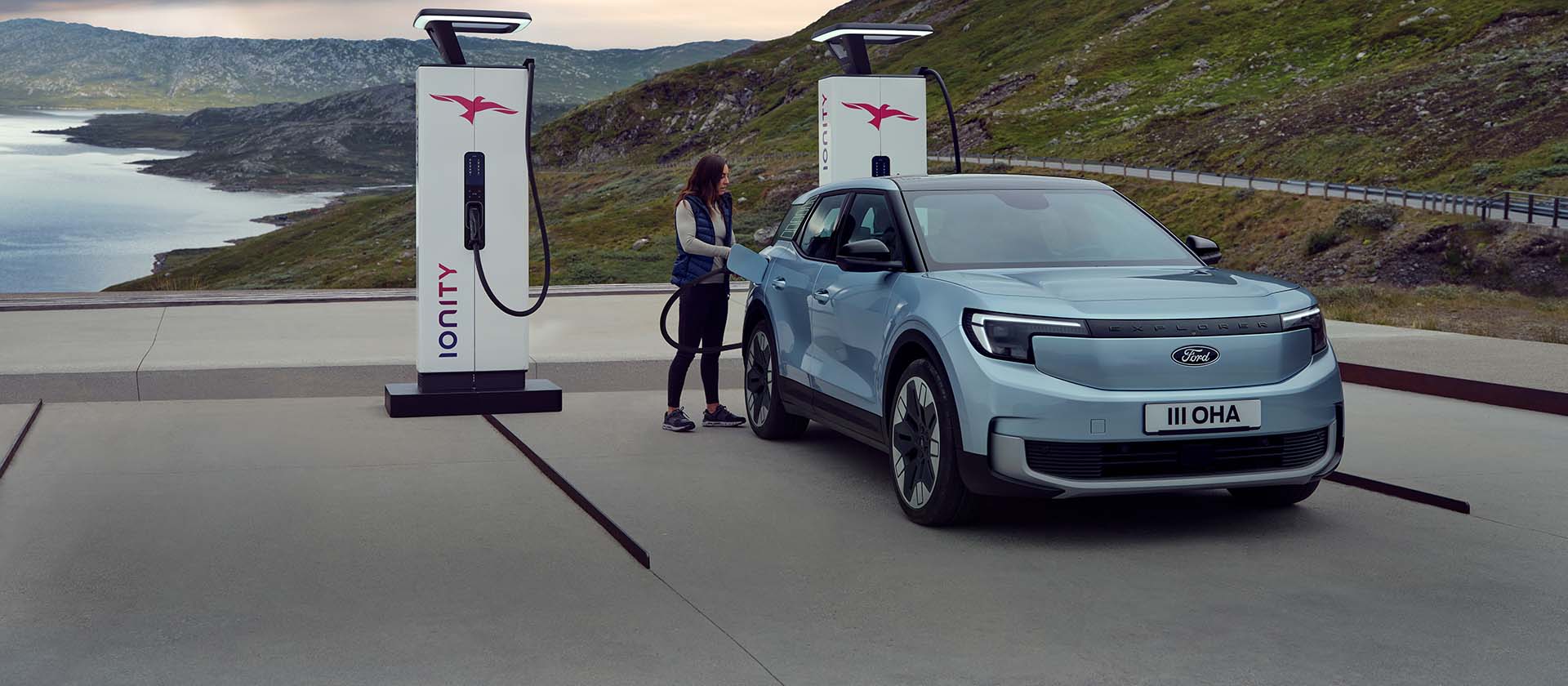 Nainen kytkee sinisen, täyssähköisen Ford Explorerin kaapelilla julkiseen latausasemaan. Auto on pysäköity vuorten ja veden äärelle.