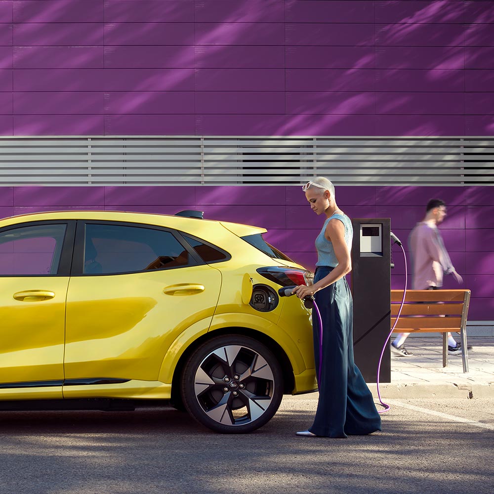 Kuljettaja kytkemässä hopeanvärisen Ford Mustang Mach-E:nsa lataukseen julkisella latausasemalla.