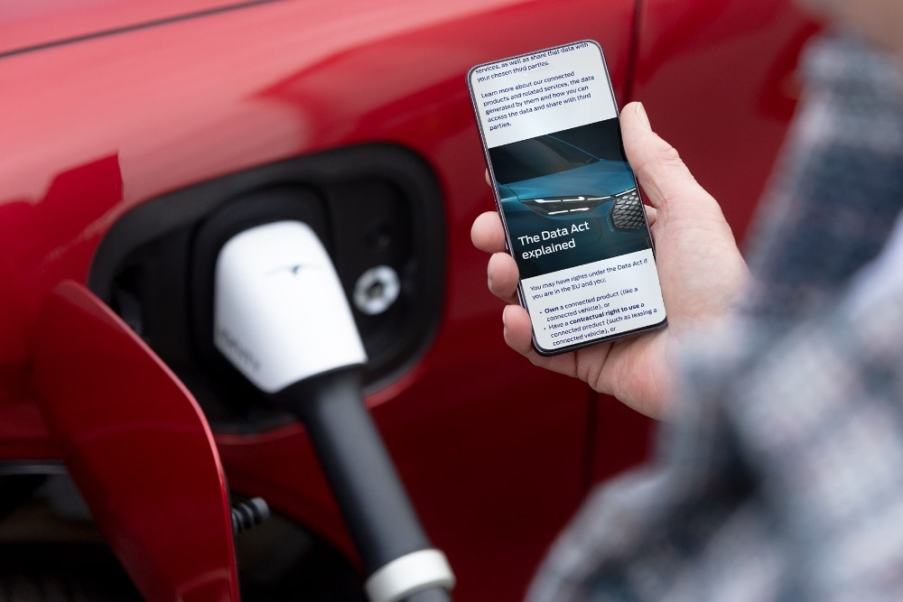 Henkilö katsoo älypuhelintaan; taustalla latautuu punainen sähköauto.
