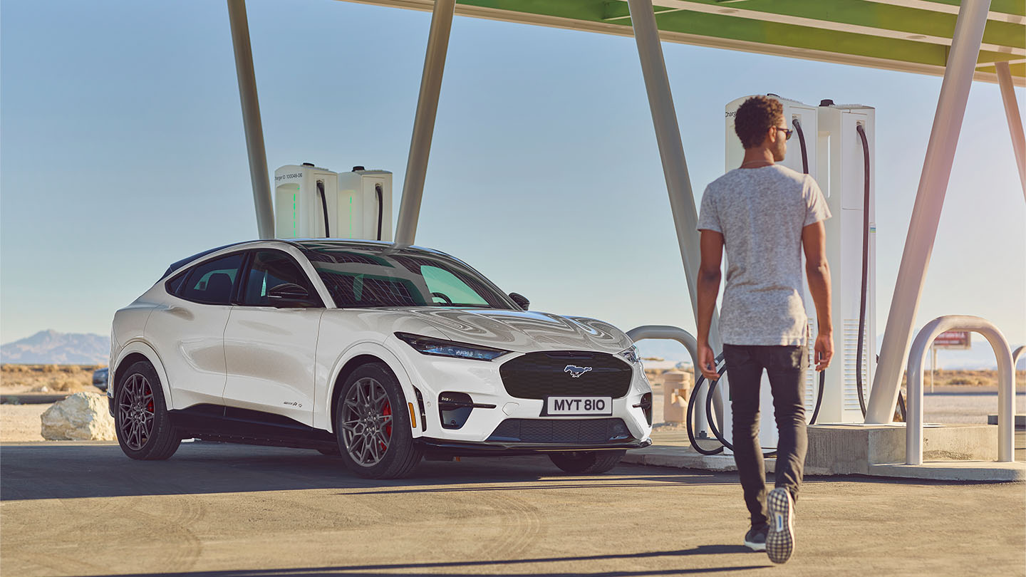 Valkoinen Ford Mustang Mach-E pysäköitynä julkiselle latausasemalle. Mies valkoisessa T-paidassa kävelee kohti autoa.