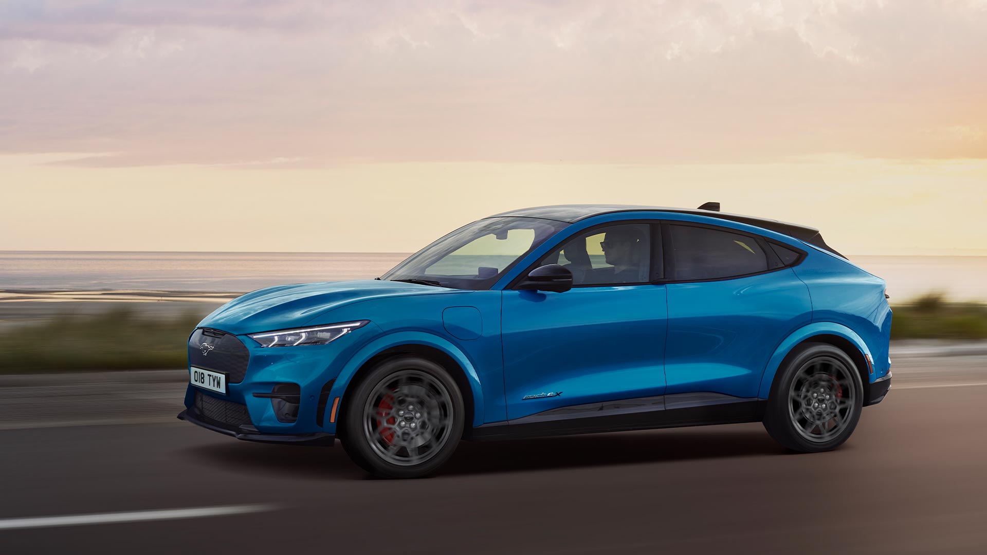 Sivusta kuvattu sininen sähköinen Ford Mustang Mach-E, joka ajaa rannikkoa reunustavalla tiellä iltahämärässä.