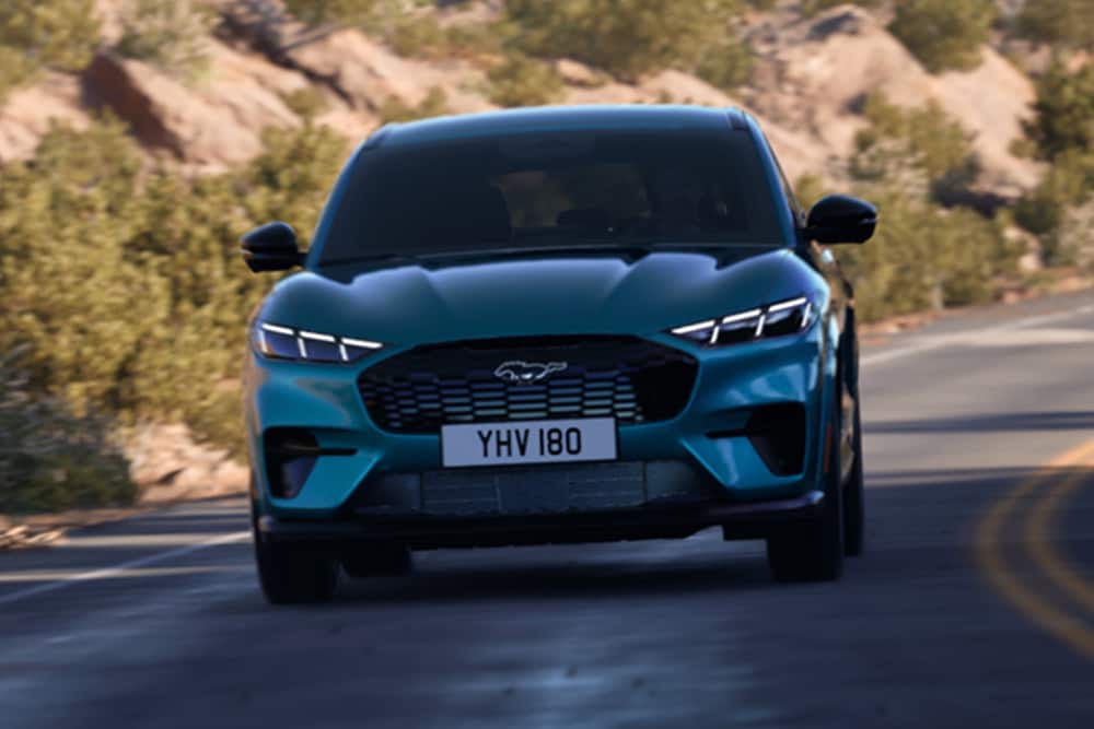 Sininen Ford Mustang Mach-E edestä kuvattuna ajamassa vuoristotiellä.