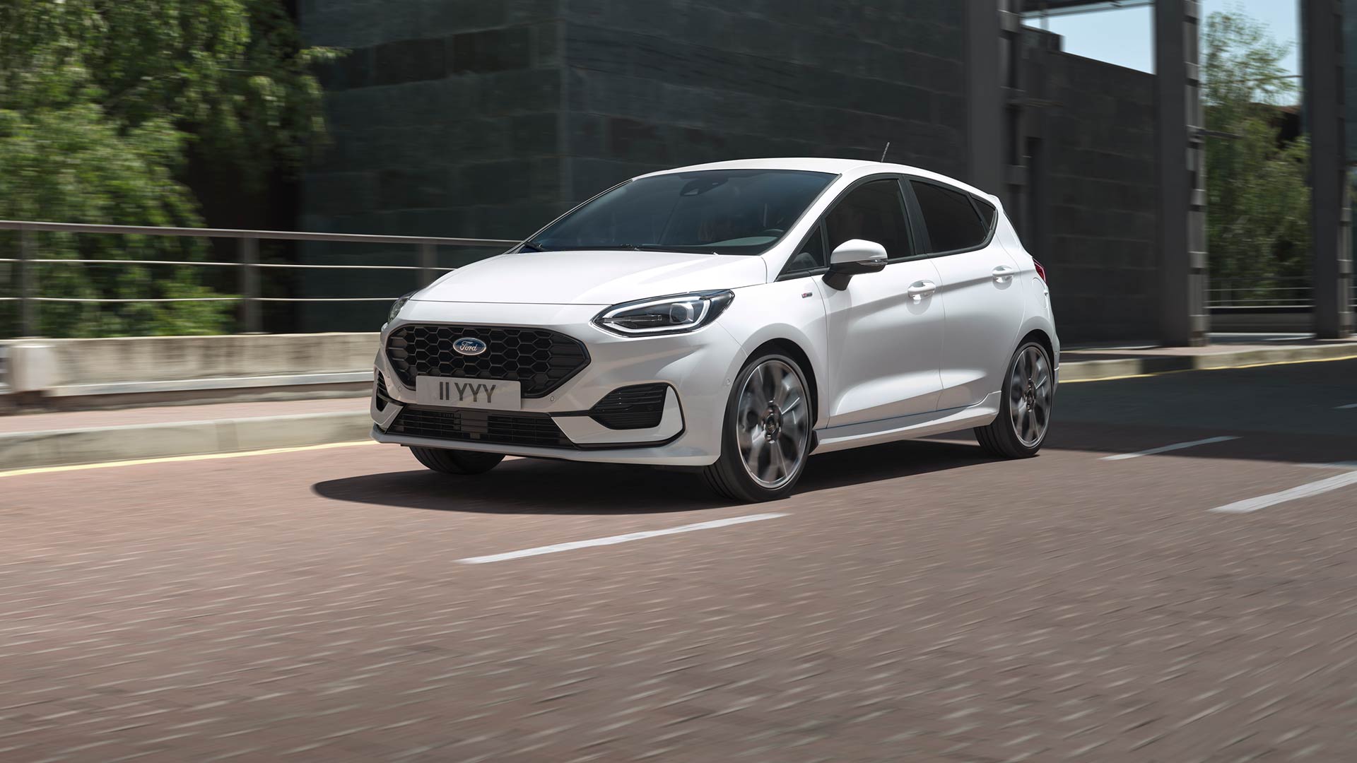 Valkoinen Ford Fiesta ajaa kaupungin kadulla, ja taustalla on viheralue ja rakennuksia.