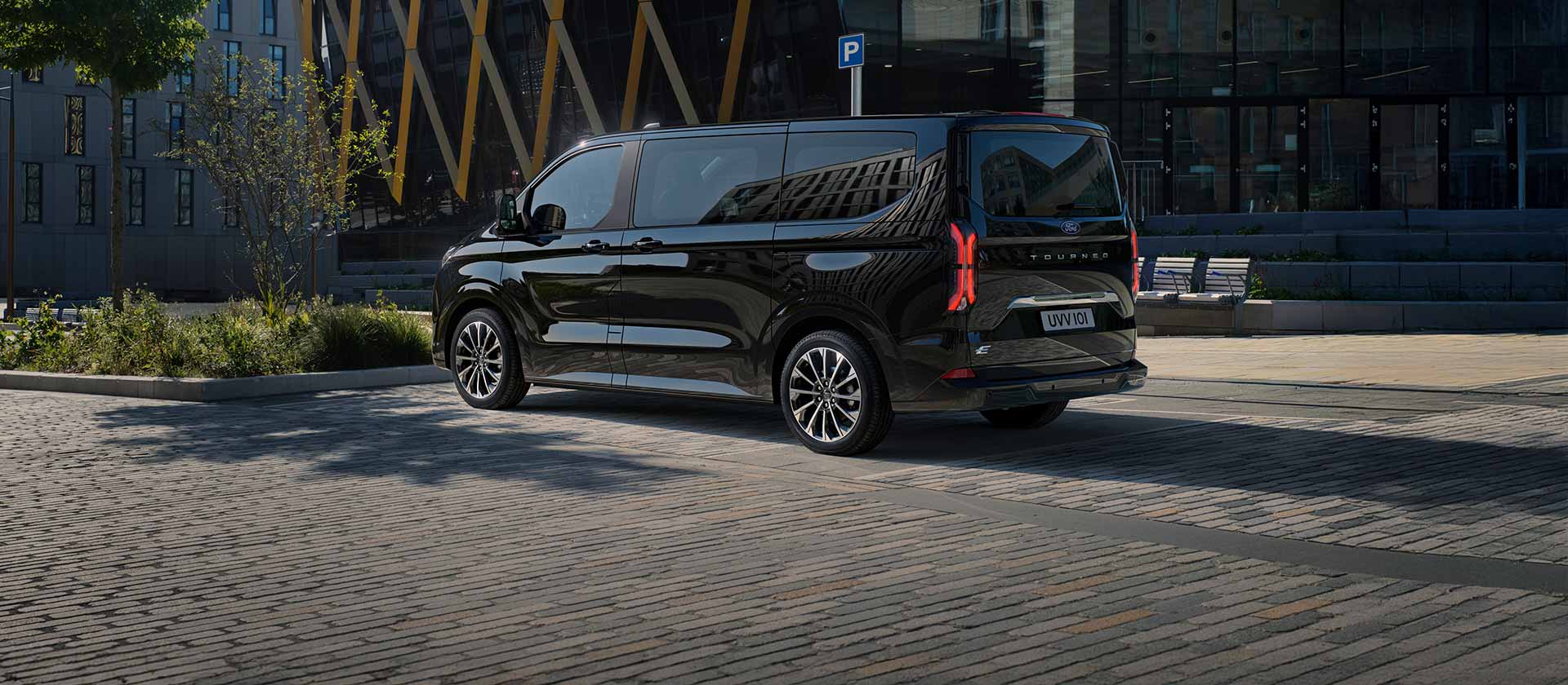 Musta Ford E-Tourneo Custom pysäköitynä modernin rakennuksen eteen ja kuvattuna takaviistosta.