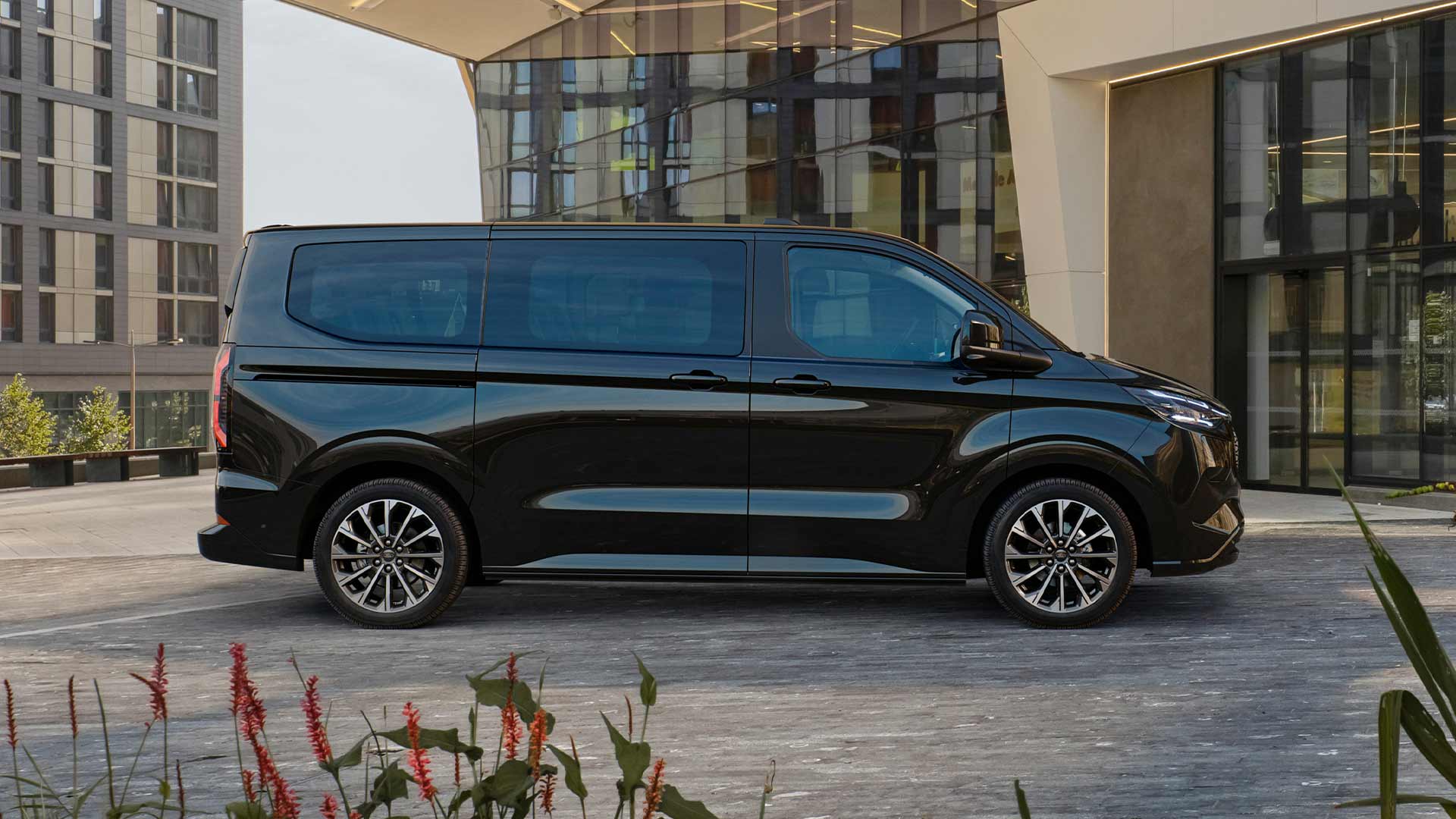 Musta Ford E-Tourneo Custom pysäköitynä modernin rakennuksen eteen.