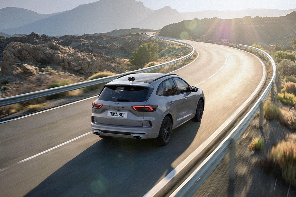 Hopeanvärinen Ford Kuga ajamassa maisemareitillä, taustalla aurinkoinen vuoristomaisema.