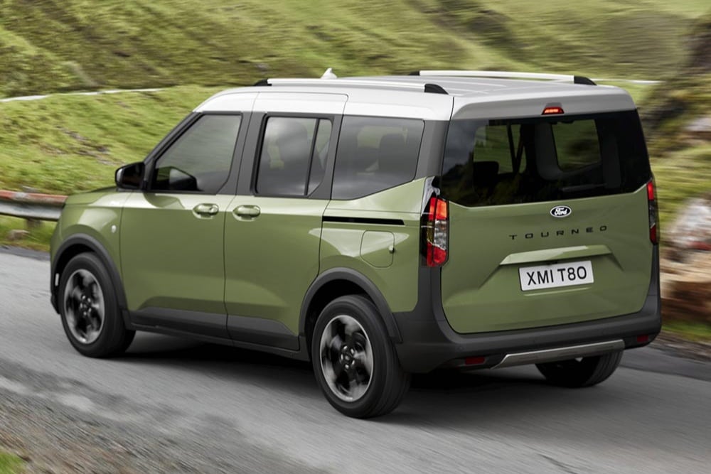 Vihreä Ford Tourneo Courier takaa viistosti kuvattuna, ajamassa mutkaisella tiellä vihreiden kukkuloiden keskellä.