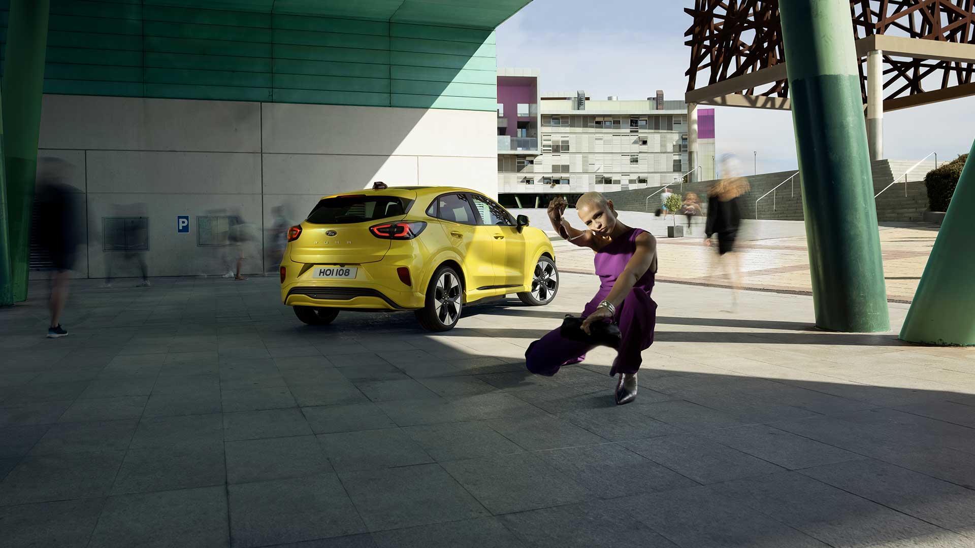 Sillan alle pysäköity keltainen sähköinen Ford Puma Gen-E® kuvattuna takaa ja vierellään violettiin pukeutunut henkilö.