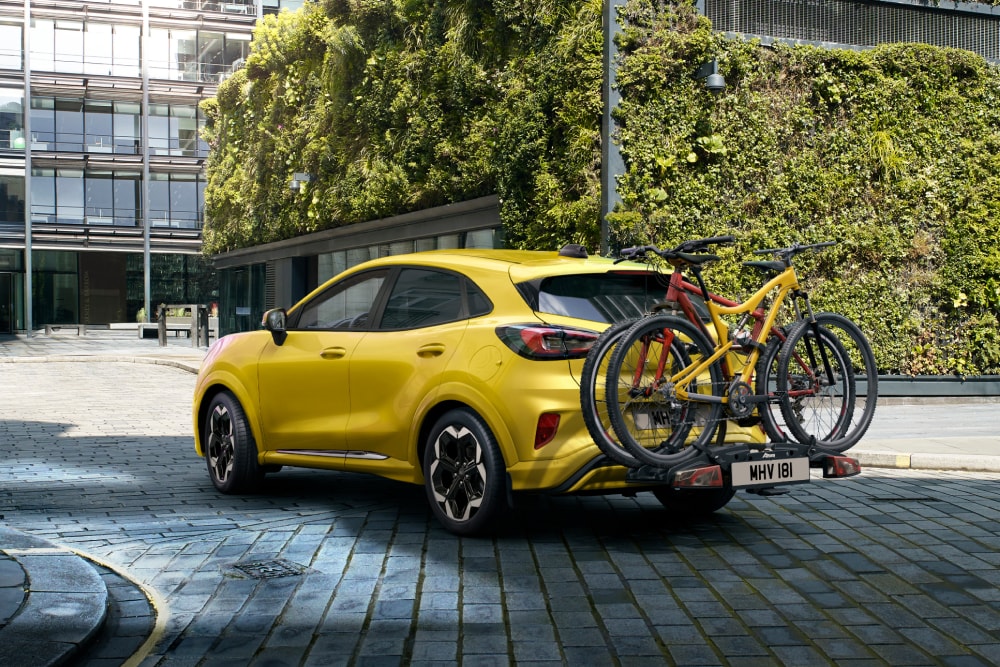 Keltainen Ford Puma Gen-E pysäköitynä kaupunkiympäristöön, kaksi polkupyörää kiinnitettynä takana olevaan pyörätelineeseen.
