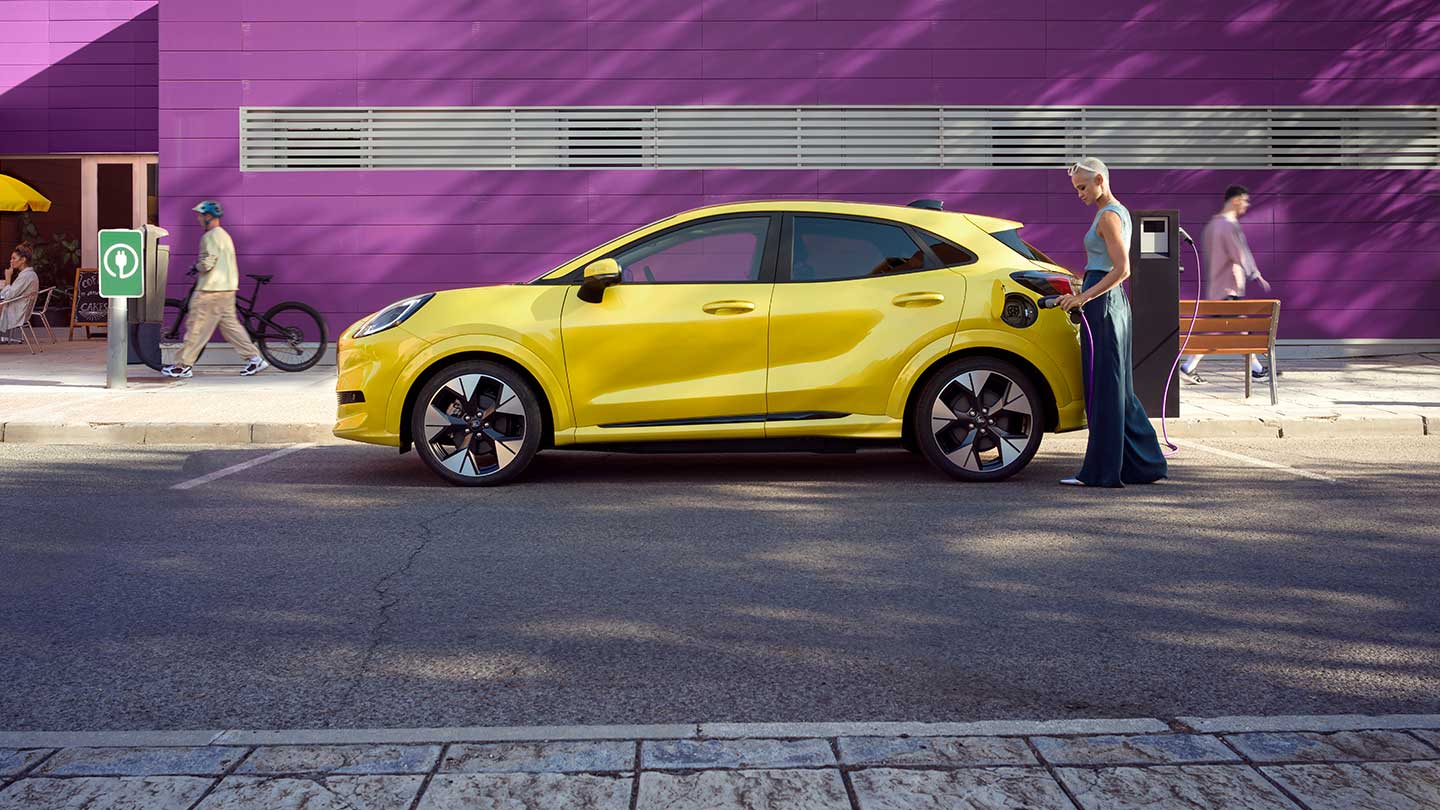 Sivunäkymä keltaisesta Ford Puma Gen-E® -sähköautosta, joka on pysäköitynä ja jota ollaan kytkemässä julkiseen latausasemaan.