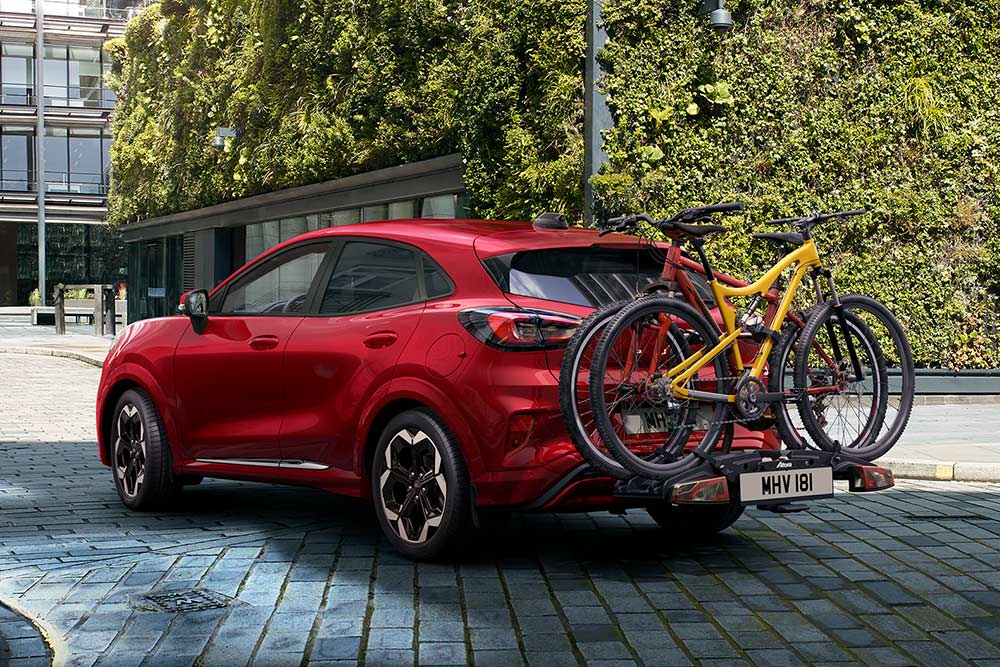 Punainen auto pysäköitynä kaupunkiympäristöön, kaksi polkupyörää kiinnitettynä takana olevaan pyörätelineeseen.