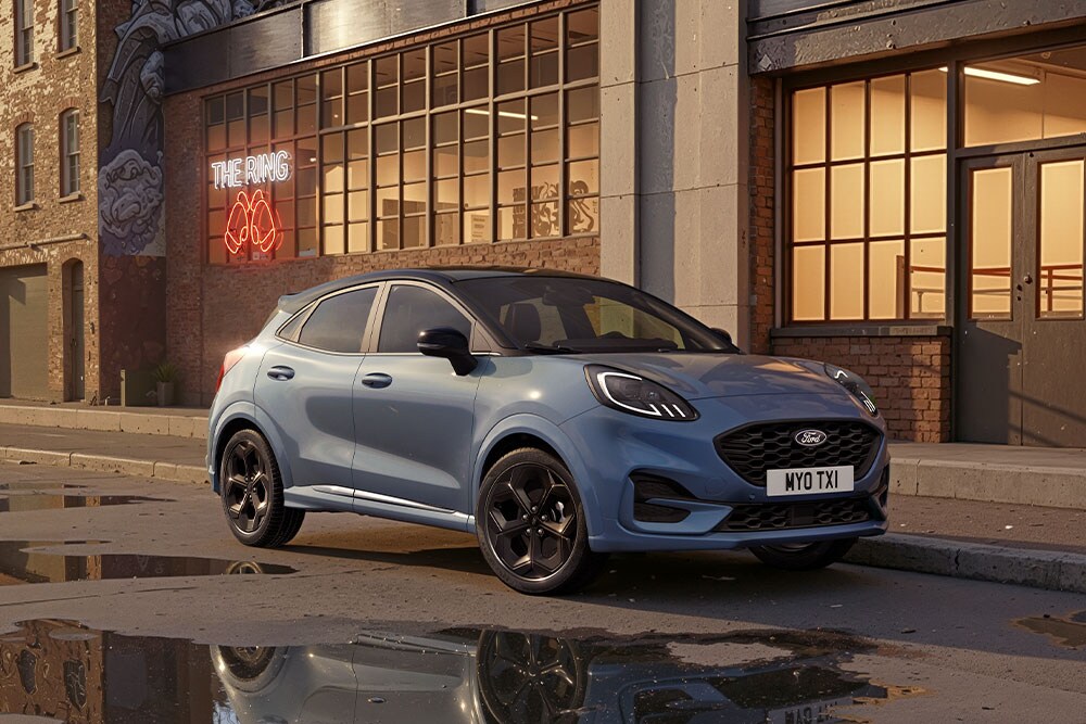 Sininen Ford Puma pysäköitynä kaupungin kadulle auringonlaskun aikaan, lätäköistä heijastuu rakennuksia.
