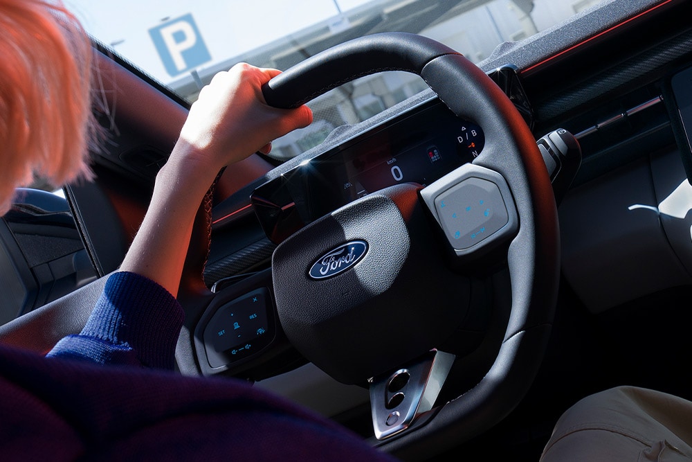 Lähikuva naisesta, joka pitelee ohjauspyörää pysäköidyssä Ford Caprissa. Kuvassa näkyy myös digitaalinen mittaristo.