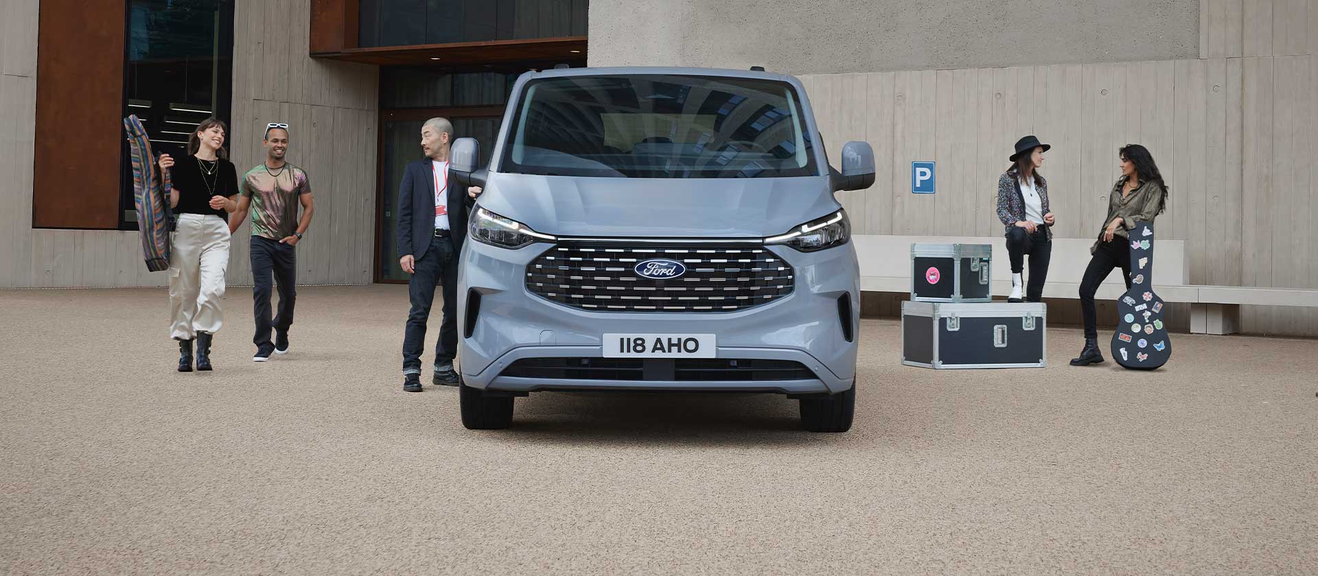 Ryhmä ihmisiä seisoo musiikkisoittimien ja -laitteiden kanssa hopeanvärisen Ford Tourneo Custom -pakettiauton lähellä.