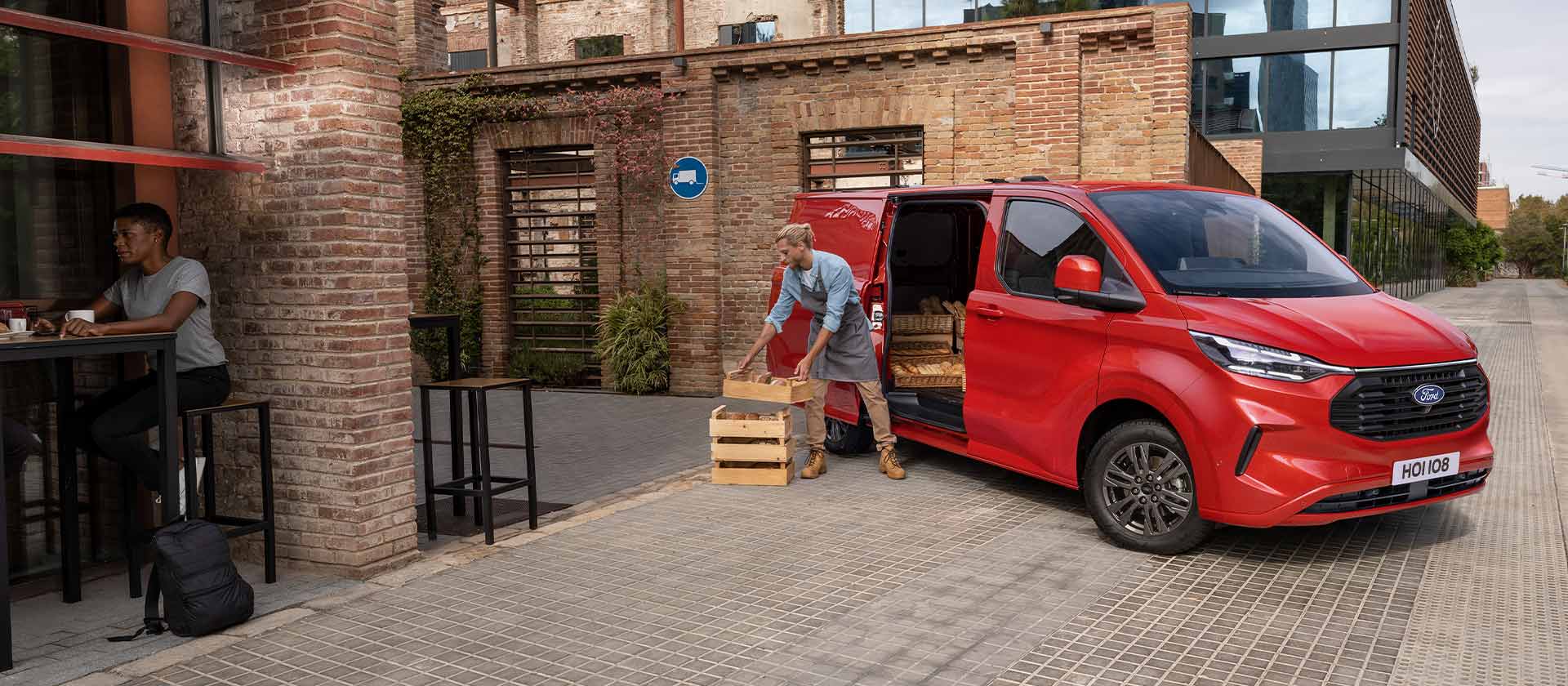 Henkilö purkaa laatikoita kahvilan eteen pysäköidystä punaisesta Ford Transit Custom -pakettiautosta. 