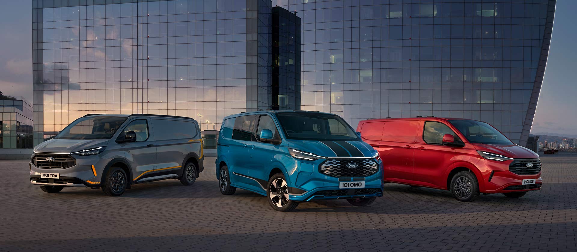 Kolme pysäköityä Ford Transit Custom -pakettiautoa: harmaa, sininen ja punainen. 