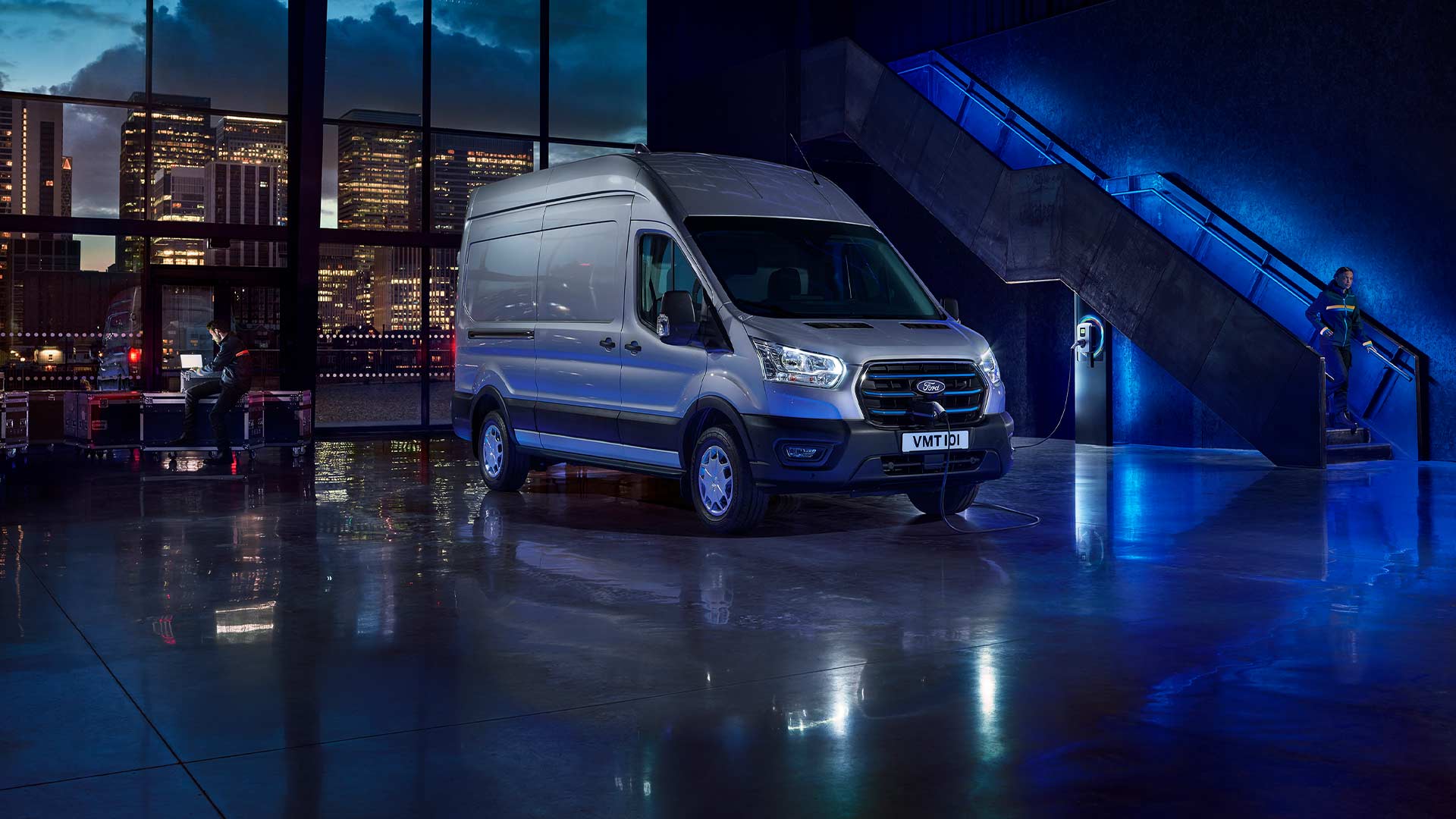 Hopeanvärinen Ford E-Transit -sähköpakettiauto pysäköitynä sisälle; isoista ikkunoista näkyy kaupungin siluetti. 