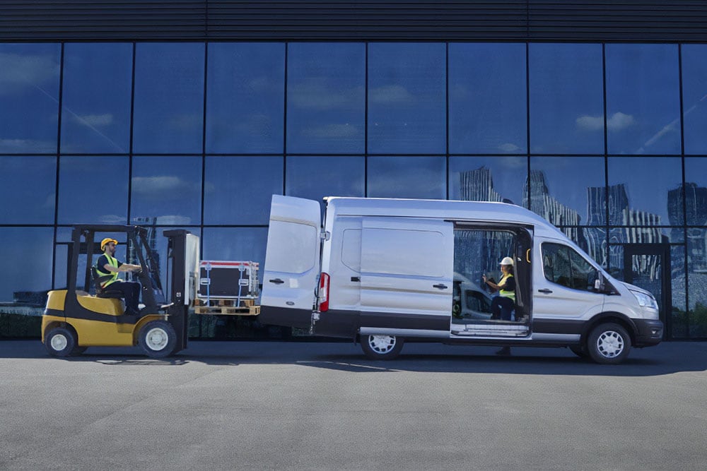 Kaksi henkilöä lastaamassa kuormaa hopeanväriseen Ford Transit -pakettiautoon haarukkatrukilla.