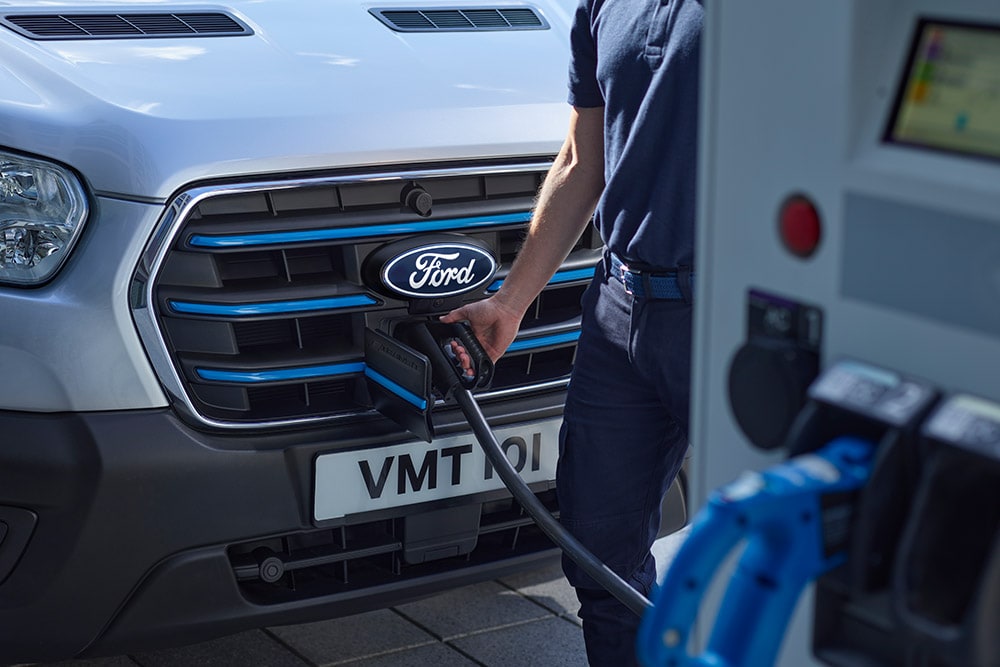 Lähikuva henkilöstä, joka yhdistää latauskaapelia hopeanväriseen Ford E-Transit -sähköpakettiautoon.