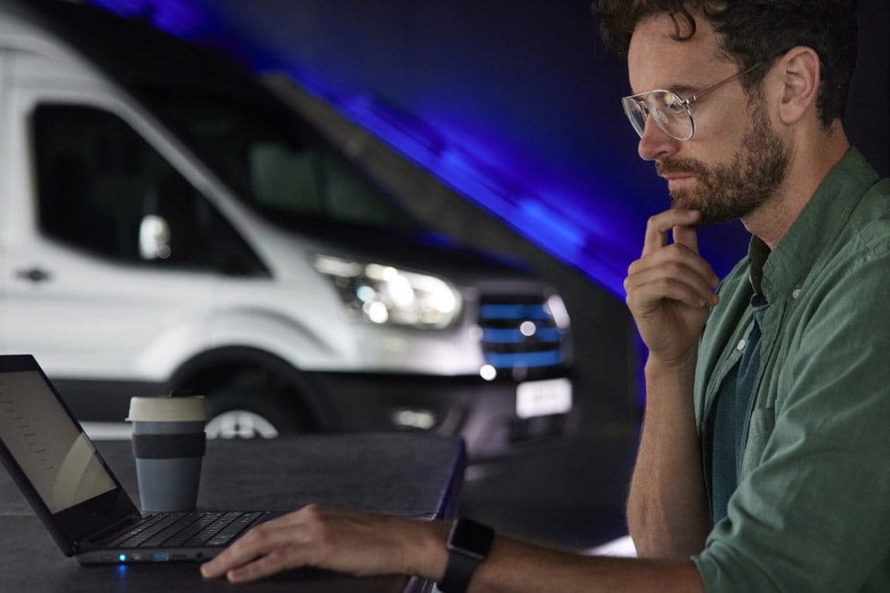 Mies työskentelemässä kannettavalla tietokoneella; taustalla Ford E-Transit -pakettiauto.
