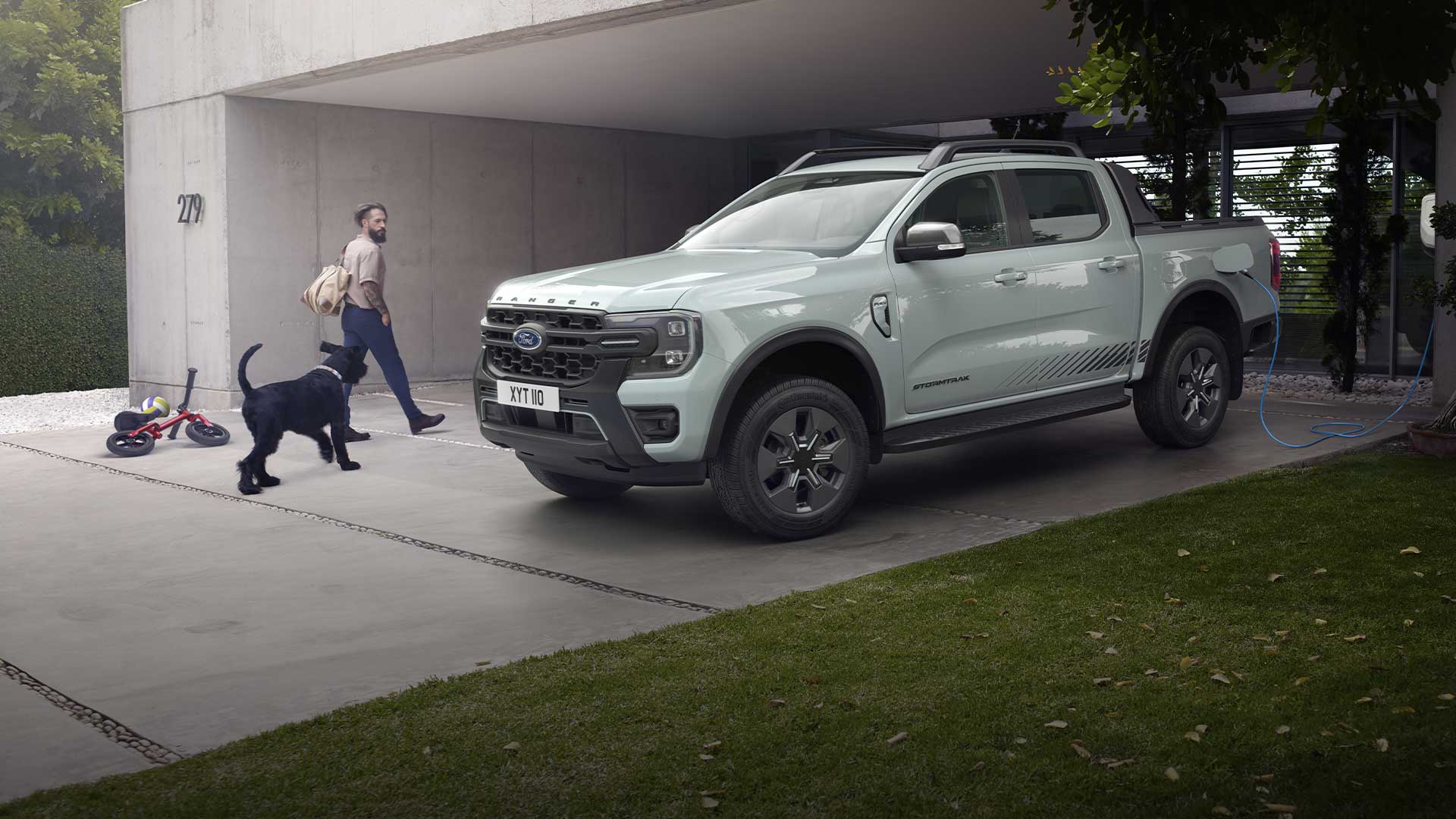 Mies ja koira kävelemässä kohti pihatiellä latauksessa olevaa harmaata Ford Ranger ladattava hybridi -avolava-autoa. 