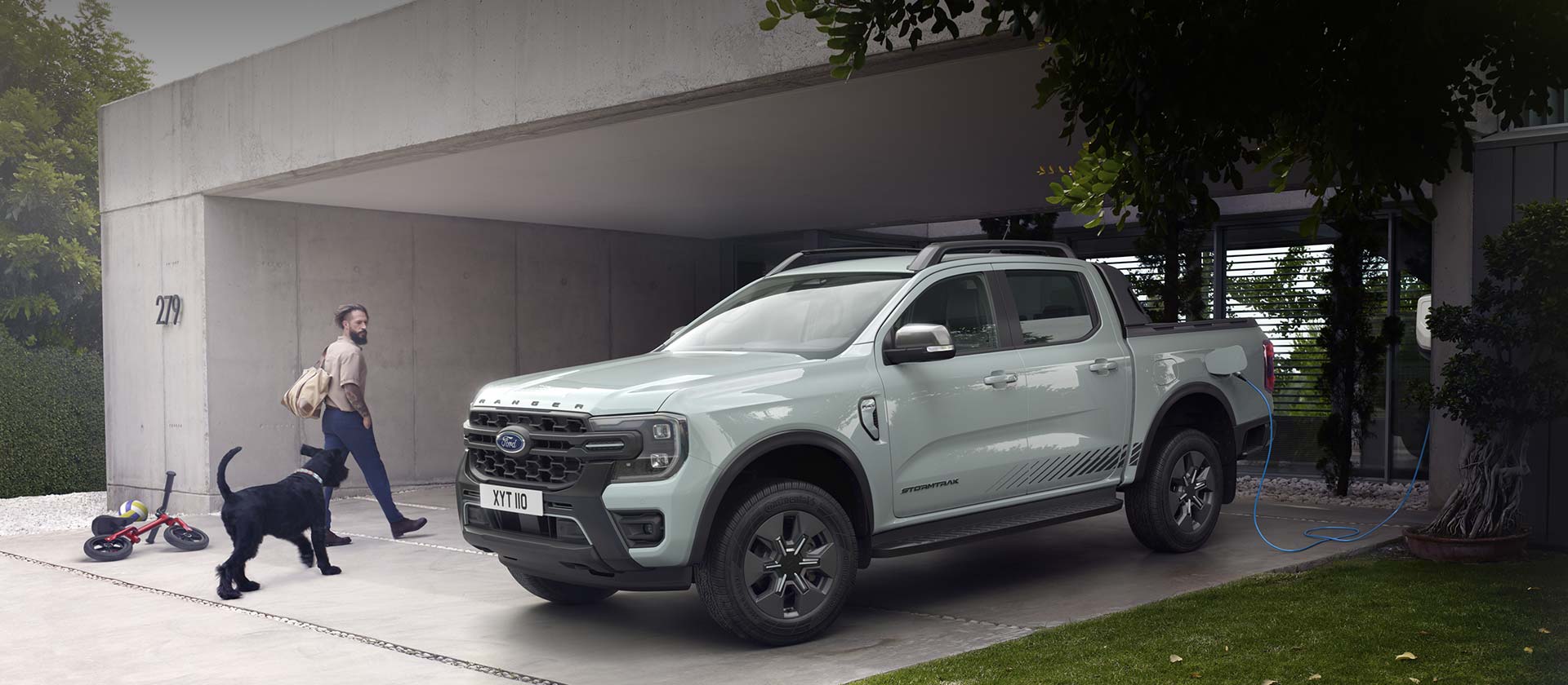 Mies ja hänen koiransa kävelevät moderniin taloon ja ohittavat pysäköidyn ja latauksessa olevan Ford Ranger ladattavan hybridin. 