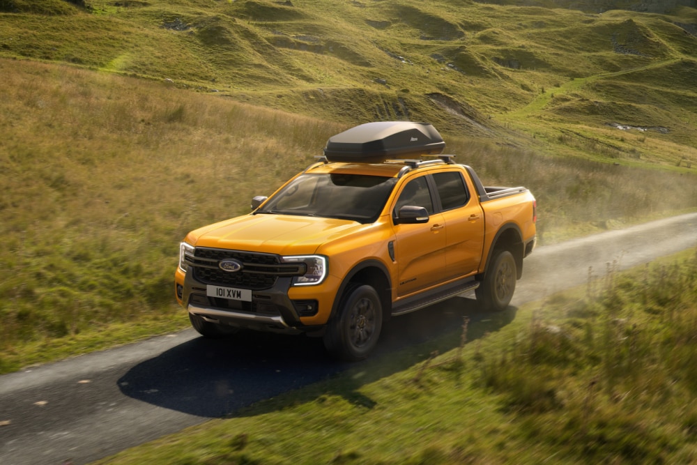 Keltainen Ford Ranger, jonka katolla oleviin taakkatelineen poikittaistankoihin on kiinnitetty kattokuljetuslaatikko, on ajamassa maaseudulla soratietä pitkin.
