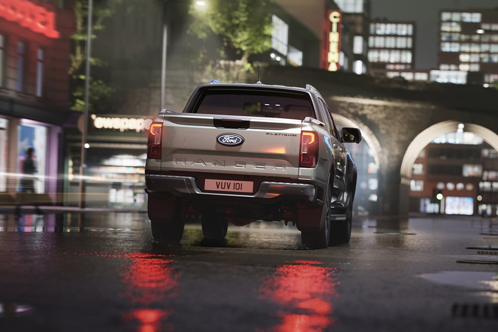 Takaapäin kuvattu hopeanvärinen Ford Ranger ajaa kaupungissa neonvalojen valaisemalla kadulla iltamyöhällä.