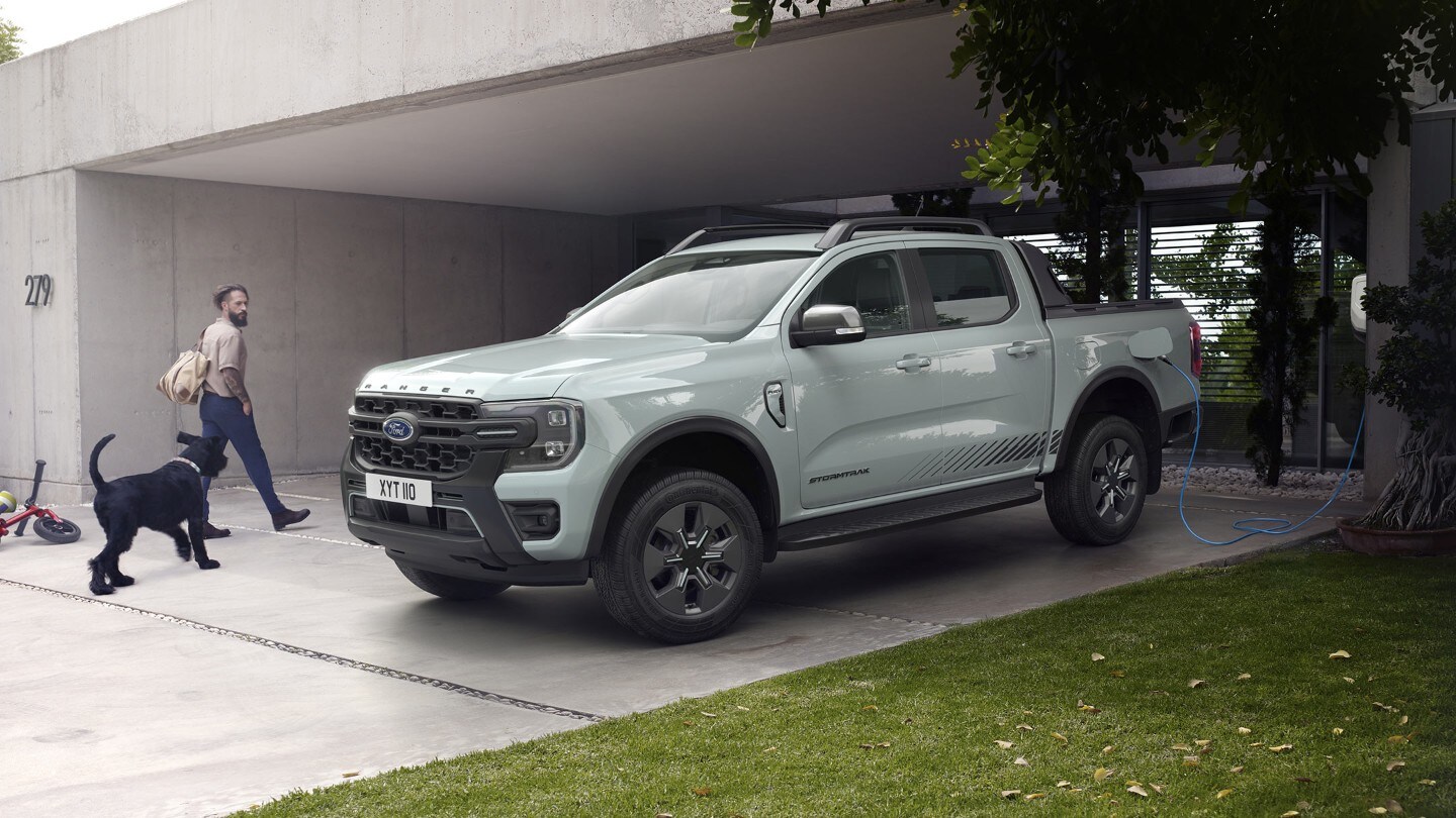 Mies ja hänen koiransa kävelevät moderniin taloon ja ohittavat pysäköidyn ja latauksessa olevan Ford Ranger ladattava hybridi pick-upin.