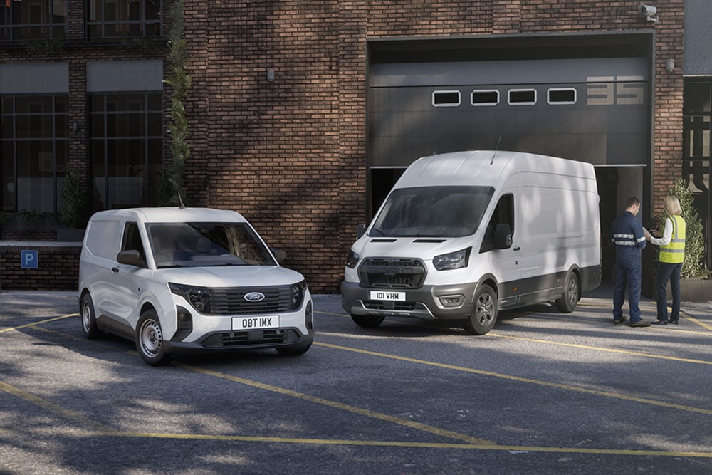 Kaksi valkoista Ford-pakettiautoa pysäköitynä modernin rakennuksen eteen; kaksi henkilöä juttelemassa autojen vieressä.