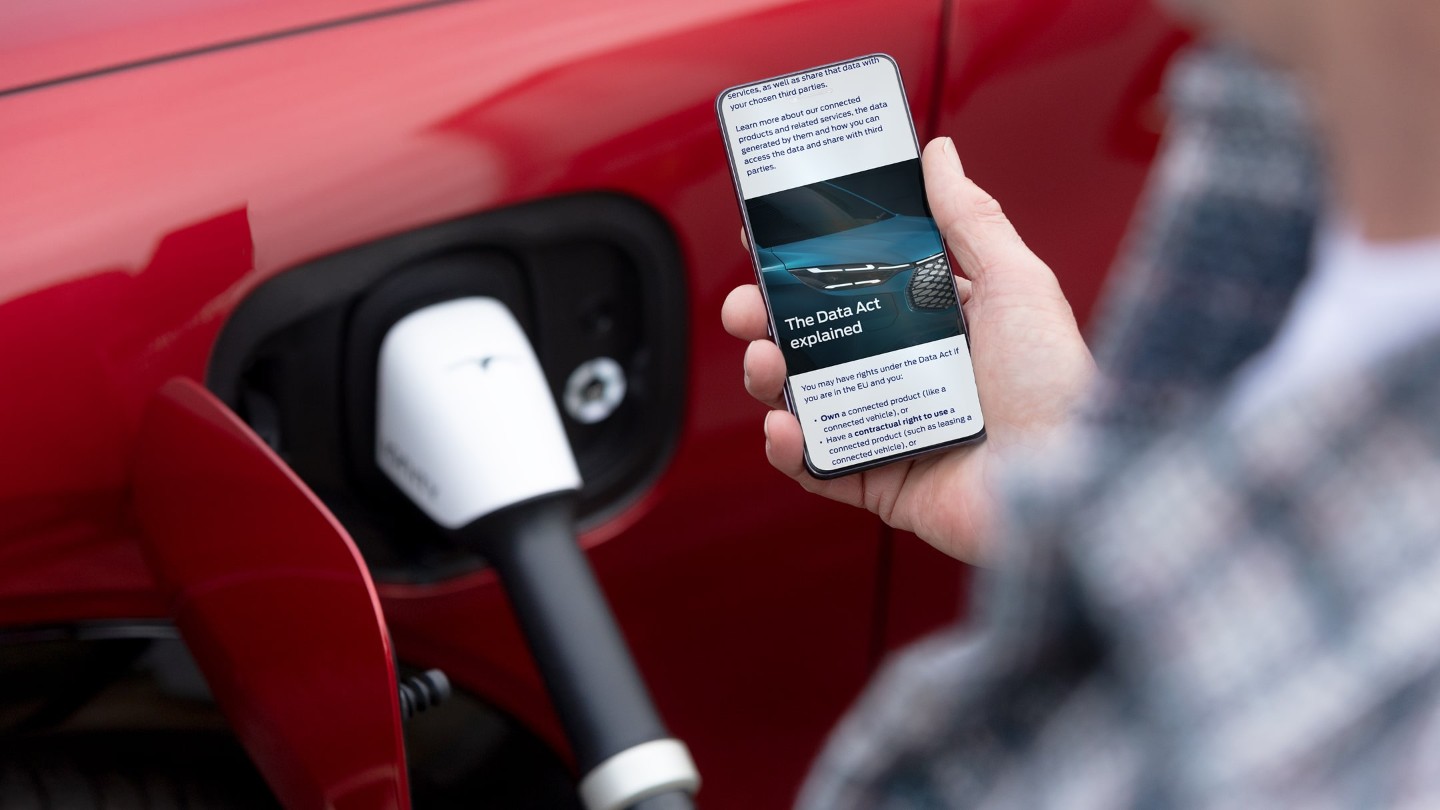 Henkilö katsoo älypuhelintaan; taustalla latautuu punainen sähköauto.