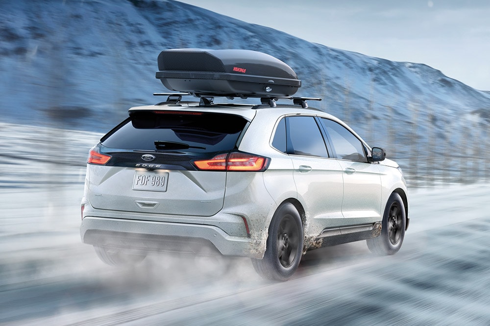 Valkoinen Ford Edge kattoboksilla ajaa lumisella vuoristotiellä, liikkeen sumennus.